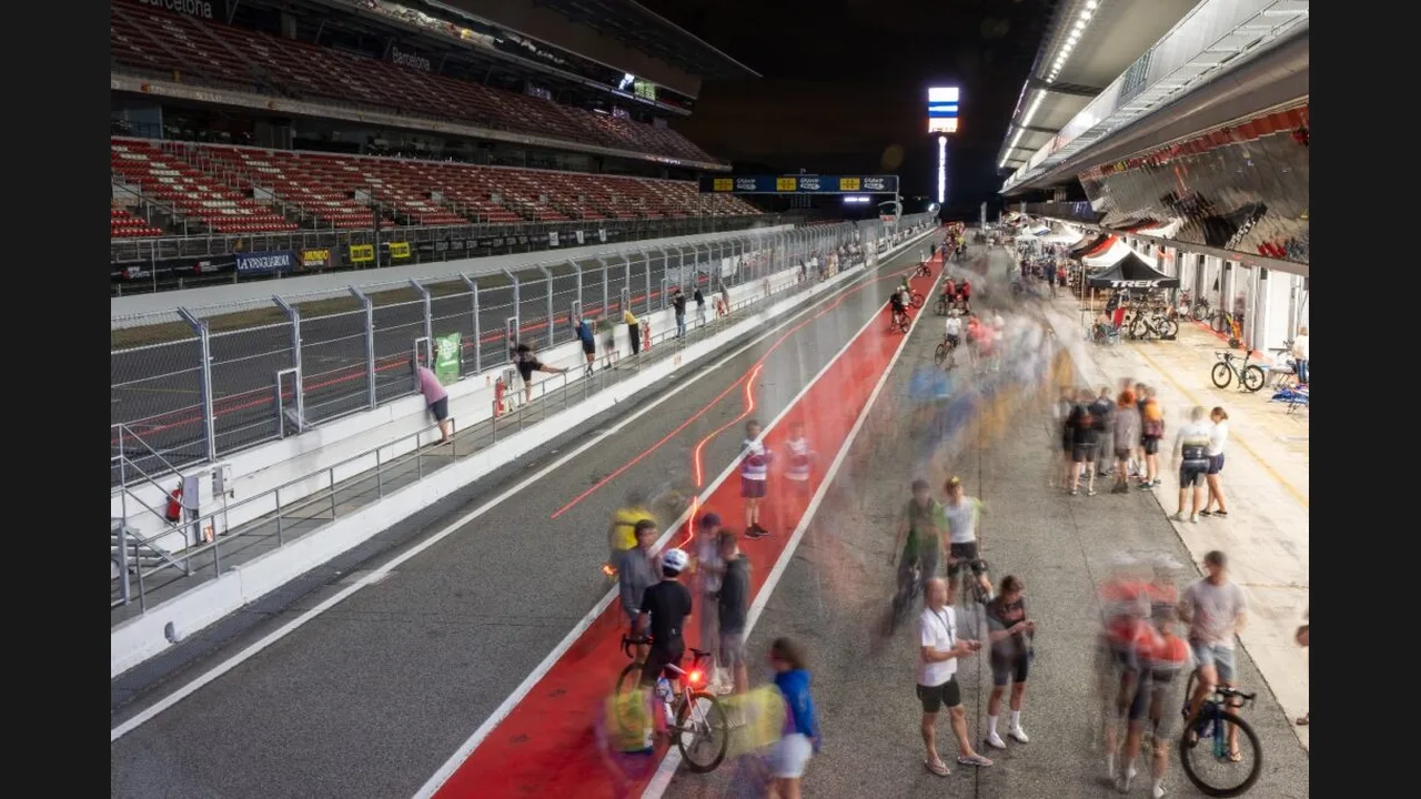 Cyclists on a race circuit at night beside a grandstand and red-and-white kerbs