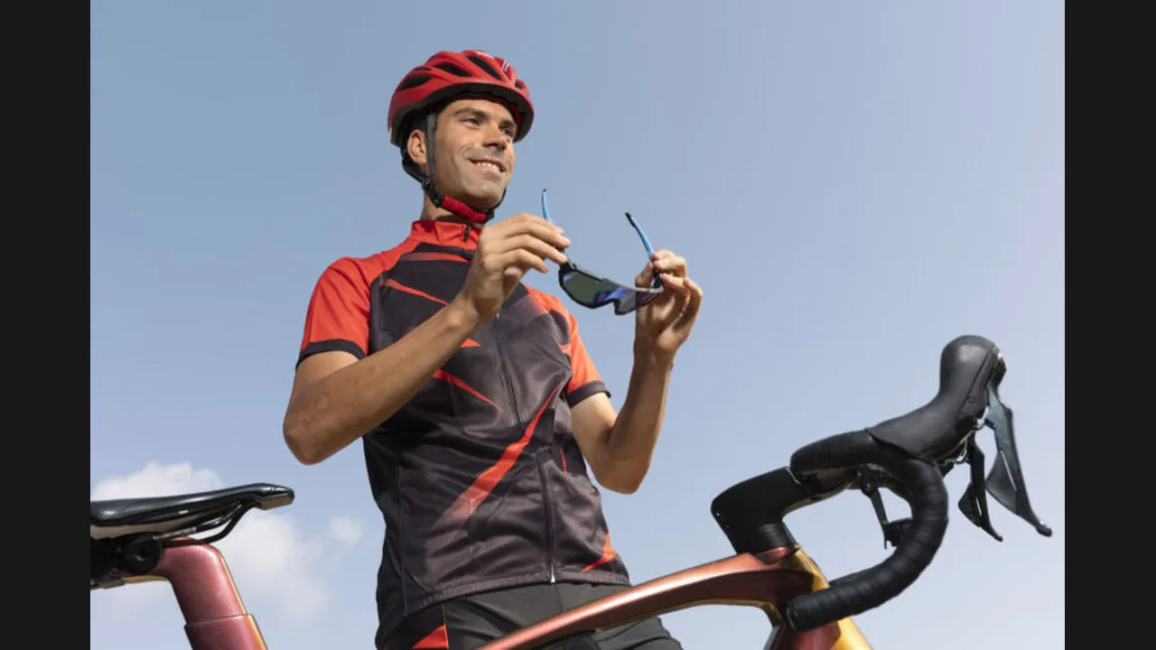 A cyclist in a red helmet and red-and-black jersey holding sunglasses beside his bicycle